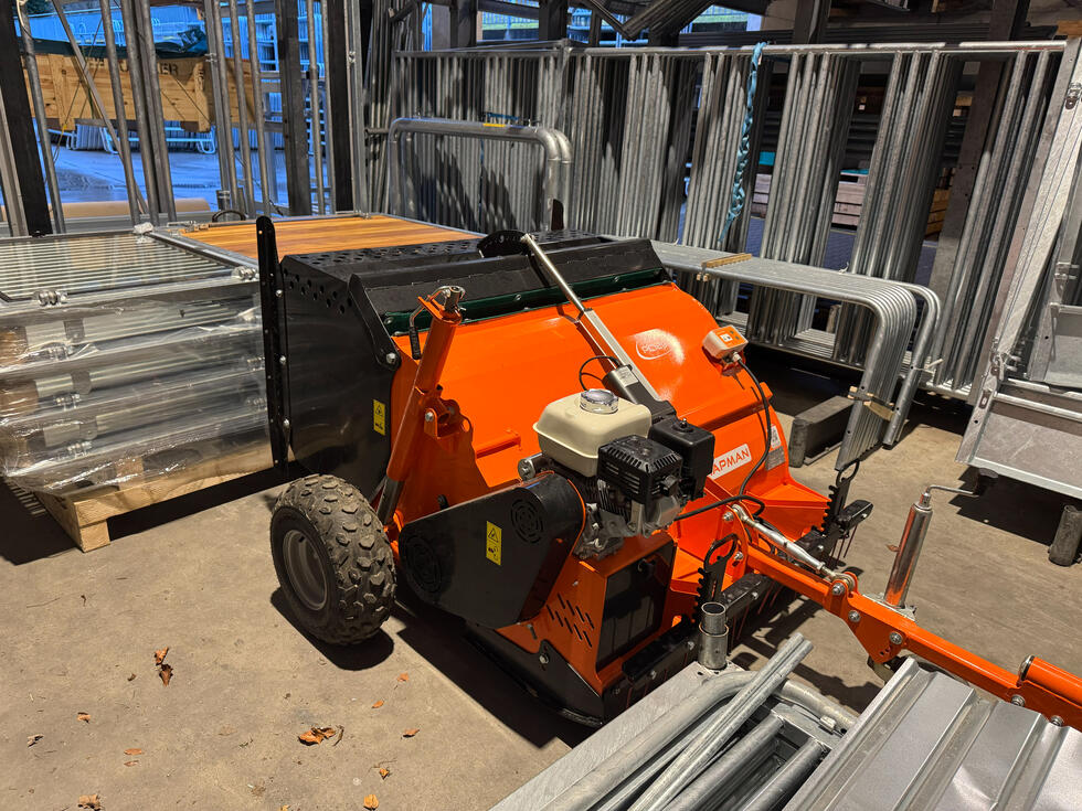 Eine Schubkarre mit Metallmulde, rot lackiertem Rohrgestänge, daraus zwei schwarz gummierte Lenkgriffe, metallener Motorkasten unterlegt, zwei festen Rädern vorne, mit Ackerstollen bereift, einem schwenkbaren Rad hinten, auf einer Baustelle, eine Person an den Griffen, wird von einem Bagger befüllt