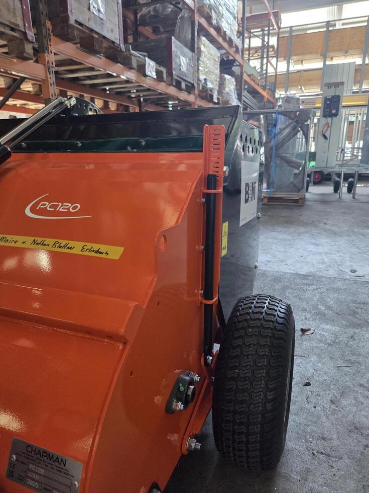 Eine Schubkarre mit Metallmulde, rot lackiertem Rohrgestänge, daraus zwei schwarz gummierte Lenkgriffe, metallener Motorkasten unterlegt, zwei festen Rädern vorne, mit Ackerstollen bereift, einem schwenkbaren Rad hinten, auf einer Baustelle, eine Person an den Griffen, wird von einem Bagger befüllt