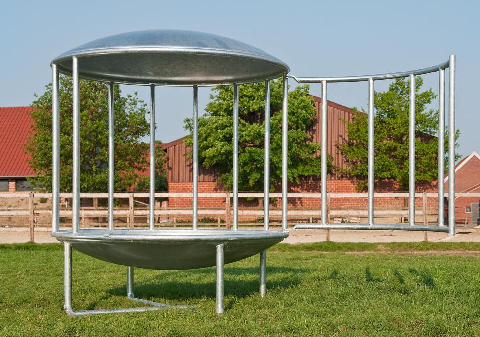 Eine Rundraufein zylindrischer Form mit einem gewölbten, verzinkten Deckel und Boden, dazwischen senkrechte Verstrebungen, resultierend in fünfzehn Fressplätzen, unterliegend zwei gekreuzte Stangen als Gestell, geöffnet und auf einer Weide vor einem Hof stehend