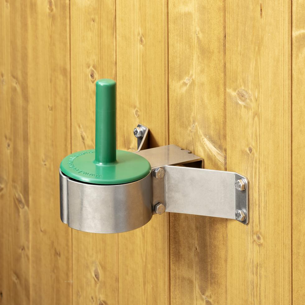 In metallischer, dreibeiniger Lecksteinhalter, eingesteckt und montiert an Flacheisen, am Ende der drei Beine befindet sich eine Rundung im Flacheisen, auf welcher ein grüner Kunststoffstab auf einer Platte befestigt ist, verschraubt an Holzwand, eine Person hängt den dreibeinigen Halter aus der Montageplatte aus