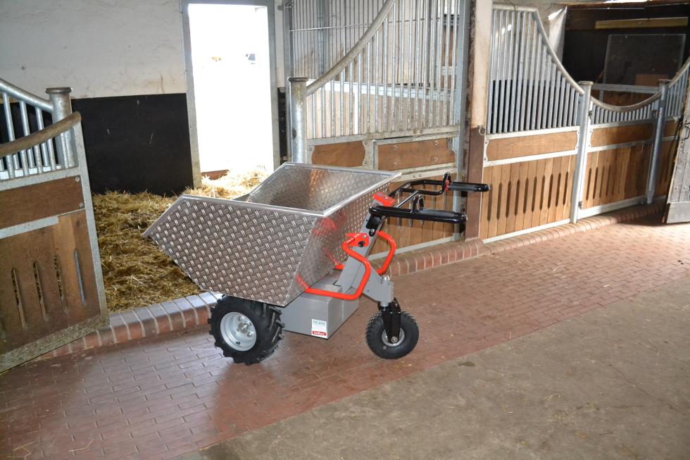 Eine Schubkarre mit Metallmulde, rot lackiertem Rohrgestänge, daraus zwei schwarz gummierte Lenkgriffe, metallener Motorkasten unterlegt, zwei festen Rädern vorne, mit Ackerstollen bereift, einem schwenkbaren Rad hinten, auf einer Baustelle, eine Person an den Griffen, wird von einem Bagger befüllt