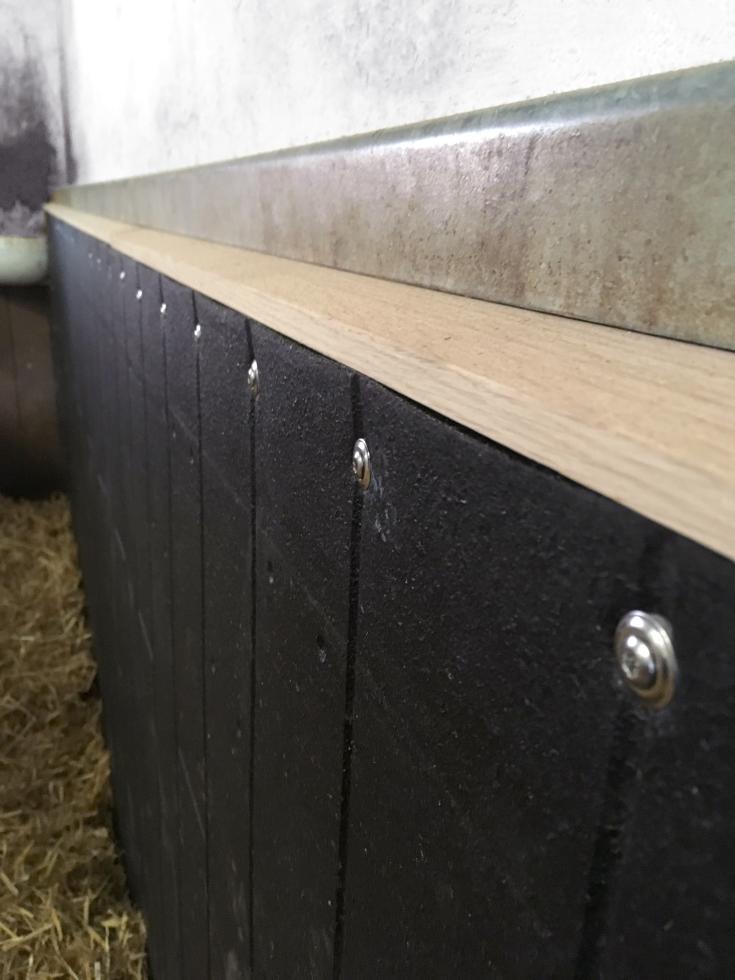 Hängende, schwarze Gummimatten in Holzbohlenoptik, festgeschraubt auf einer Holzleiste, an einer Wand, in einem Stall, schräg seitliche Detailansicht der Leiste, sowie deren Flucht