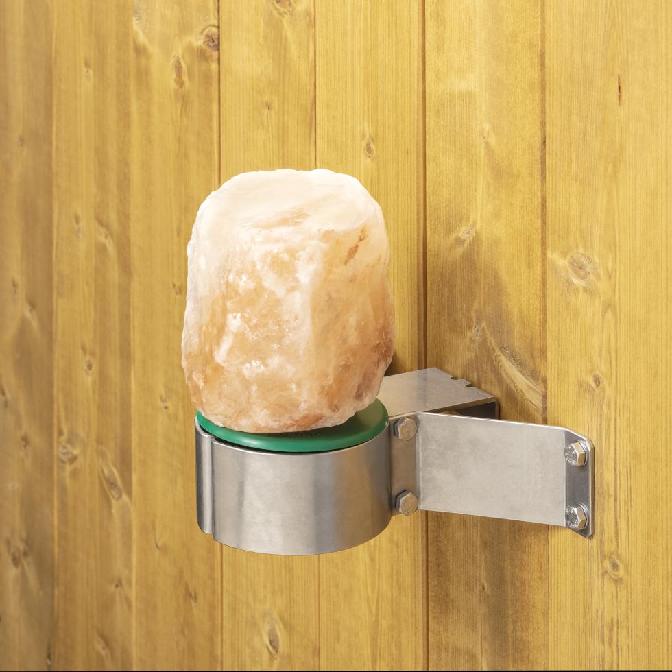 In metallischer, dreibeiniger Lecksteinhalter, eingesteckt und montiert an Flacheisen, am Ende der drei Beine befindet sich eine Rundung im Flacheisen, auf welcher ein grüner Kunststoffstab auf einer Platte befestigt ist, verschraubt an Holzwand, eine Person hängt den dreibeinigen Halter aus der Montageplatte aus