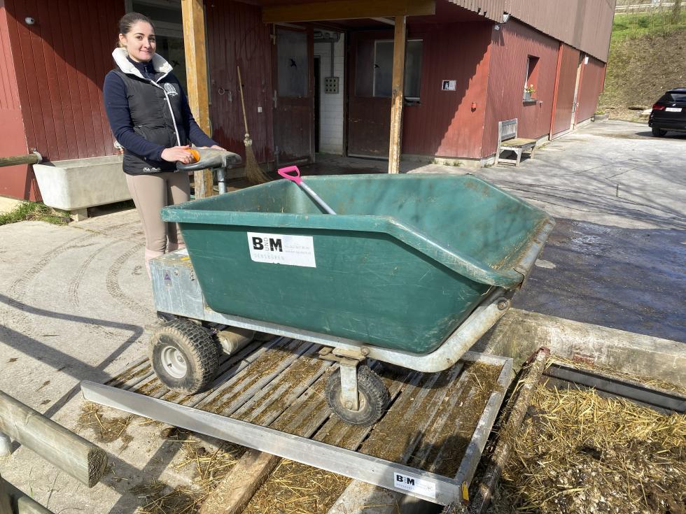 Eine grüne Kunststoffschubkarre mit vier Rädern und einem Trittbrett, hinter der Mulde befindet sich die Steuerungseinheit auf einem Vierkant mit schwarzen Griffen und orangem Halter. Das Gestell ist aus Metall, die Reifen schwarz. Eine Person fährt auf einem Trittbrett stehend eine Rampe an einem Miststock hoch