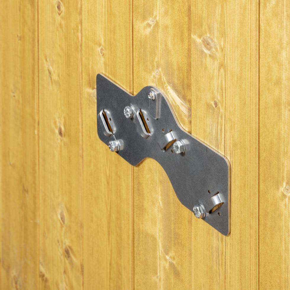 Flacheisen mit wellenartiger Ausbauchung in der Mitte und vier regelmässig angeordneter Haltelaschen für den dreibeinigen Halter, sowie vier Schrauben und oben zentral dem Sicherungshebel, montiert an Holzwand