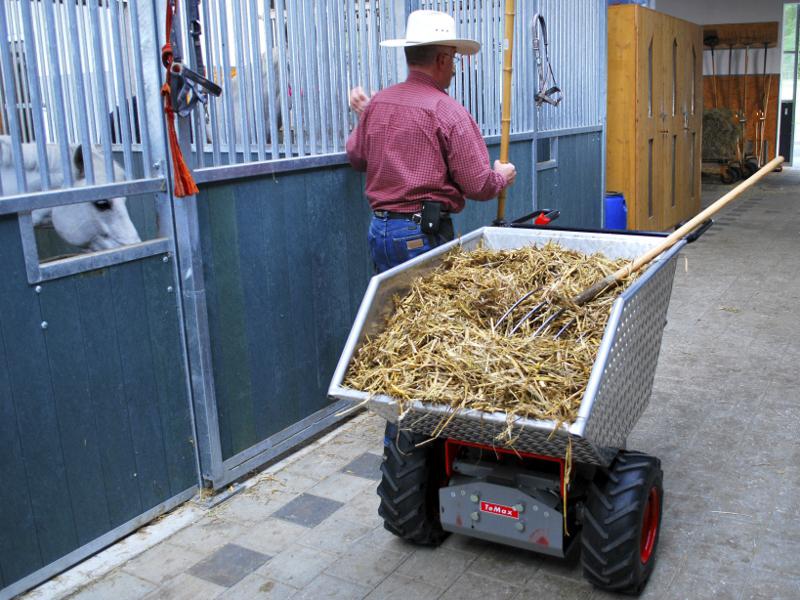 Eine Schubkarre mit Metallmulde, rot lackiertem Rohrgestänge, daraus zwei schwarz gummierte Lenkgriffe, metallener Motorkasten unterlegt, zwei festen Rädern vorne, mit Ackerstollen bereift, einem schwenkbaren Rad hinten, auf einer Baustelle, eine Person an den Griffen, wird von einem Bagger befüllt