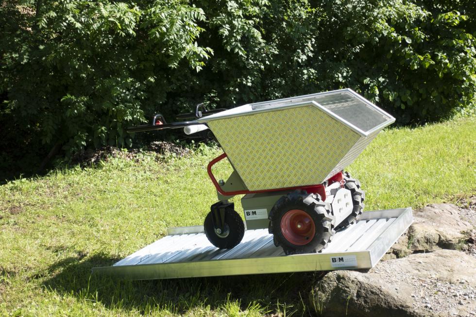 Eine Schubkarre mit Metallmulde, rot lackiertem Rohrgestänge, daraus zwei schwarz gummierte Lenkgriffe, metallener Motorkasten unterlegt, zwei festen Rädern vorne, mit Ackerstollen bereift, einem schwenkbaren Rad hinten, auf einer Baustelle, eine Person an den Griffen, wird von einem Bagger befüllt