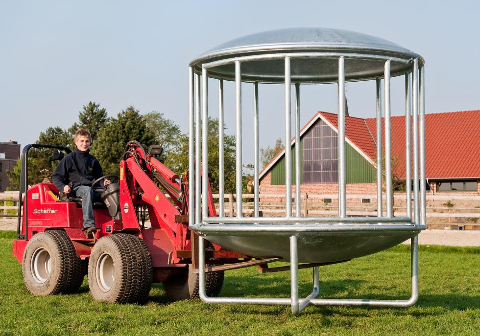 Eine Rundraufein zylindrischer Form mit einem gewölbten, verzinkten Deckel und Boden, dazwischen senkrechte Verstrebungen, resultierend in fünfzehn Fressplätzen, unterliegend zwei gekreuzte Stangen als Gestell, geöffnet und auf einer Weide vor einem Hof stehend