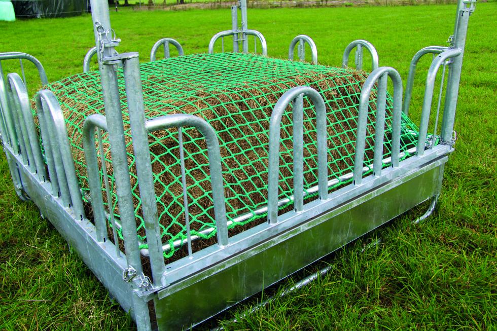 Seitliche Ansicht einer eckigen Raufe auf einer Weide, untere Hälfte Blech, obere Hälfte Rundbogengitter. In der Mitte ist ein Futterhaufen, überzogen mit einem grünen Futtersparnetz, jeweils zwei Rundbogengitter freistehend plus Eckrundbogengitter mit draufmontiertem Pfosten für Blechdach